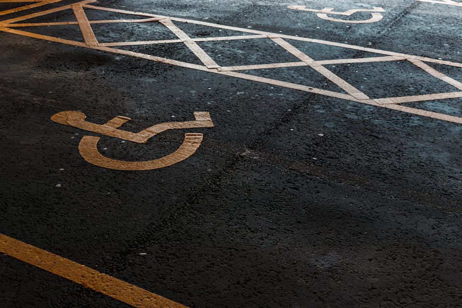 A close-up view of an asphalt parking lot surface shows a marked disabled parking space with a yellow wheelchair symbol painted on the dark, textured asphalt. The painted lines of the parking space are faded and slightly worn, with some areas appearing chipped or weathered, indicating regular use. Adjacent to the parking space is a section of diagonally striped white lines, which delineate the boundary of the accessible parking area. The background reveals more asphalt with additional parking markings, and the lighting suggests an overcast or shaded environment, providing minimal reflections or glare on the asphalt surface. The overall scene emphasizes the clarity and placement of designated disabled parking symbols within a typical urban or commercial parking lot, consistent with facilities managed by waste removal services such as Waste Removal Harringay, which often require clear access routes for service and disposal collection operations.