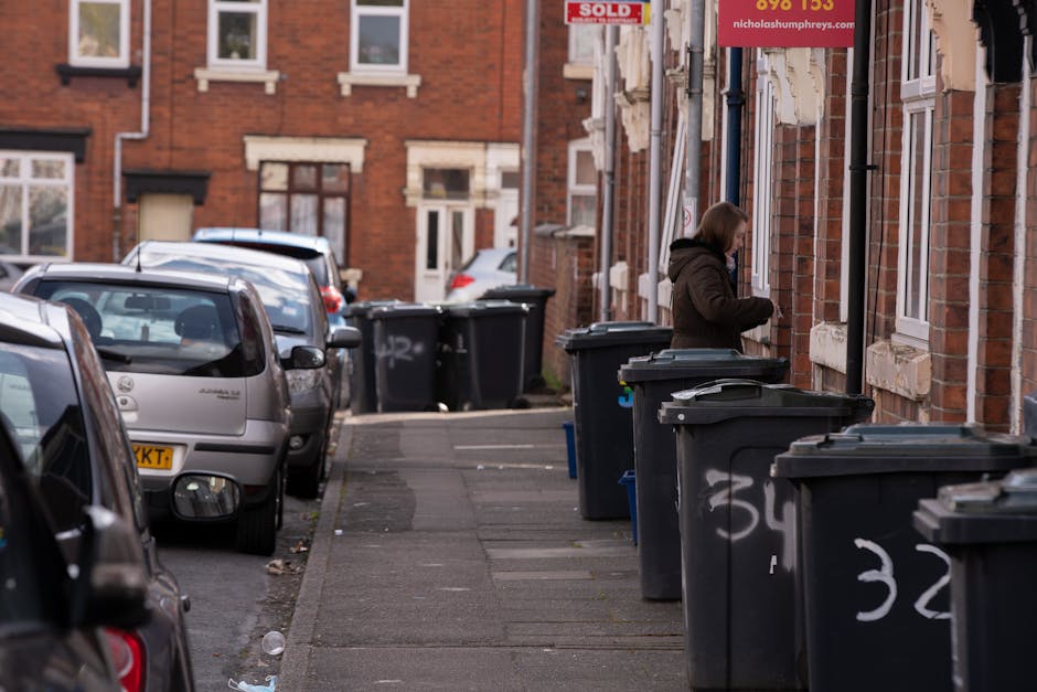A row of large black wheelie bins with closed lids lined up along a residential sidewalk adjacent to a red brick building. The bins are positioned next to each other, with some having white graffiti or markings on their sides. A woman wearing a dark coat and with shoulder-length hair is standing by one of the bins, seemingly opening or inspecting it. The background shows parked cars along the street, including a silver hatchback and other vehicles, with a narrow lead-up to the building entrances that have white-framed windows and small steps. The environment appears urban, with a calm, overcast atmosphere, and the scene reflects the typical setting for private waste disposal or on-site rubbish collection services performed by waste management companies like Waste Removal Harringay, highlighting a common approach to domestic refuse collection outside traditional municipal services.
