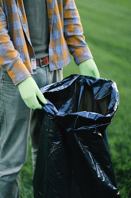 A person standing outdoors on a grassy area, wearing a plaid shirt with shades of yellow, gray, and blue over a green T-shirt, along with gray trousers secured with a brown belt. They are holding open a large, black plastic rubbish bag with both hands, which is partially filled and appears to be used for waste collection. The individual is also wearing bright green gloves, indicating they are engaged in rubbish removal or cleanup activity. The background features a well-maintained green lawn, suggesting an outdoor environment suitable for rubbish clearance, possibly as part of a garden or park tidy-up. The focus is on the person and the rubbish bag, emphasizing on-site waste collection, which may relate to independent waste handling services offered by Waste Removal Harringay, aligning with emergency or same-day rubbish removal services in the area.