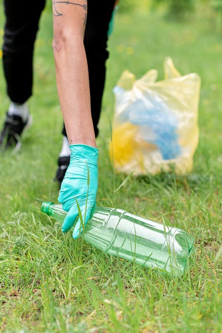 A person wearing a green disposable glove is kneeling on a grassy area, picking up a clear plastic bottle with a green cap from the ground. In the background, there is a yellow and blue plastic bag, partially filled with waste, resting on the grass. The scene appears to be outdoors, possibly in a park or garden setting, with green foliage and blurred natural surroundings. This image illustrates a clean-up activity related to rubbish removal and environmental maintenance, highlighting the importance of proper waste collection and selective debris clearing, which Waste Removal Harringay supports through private on-site clearance services. The lighting is natural daylight, casting a soft, even illumination over the scene, emphasizing the textures of the grass, plastic, and the person's arm. The overall composition reflects a careful approach to waste collection, with attention to material details such as the transparency of the plastic bottle and the smooth finish of the gloves, aligning with the practical aspects of rubbish removal and environmental responsibility.