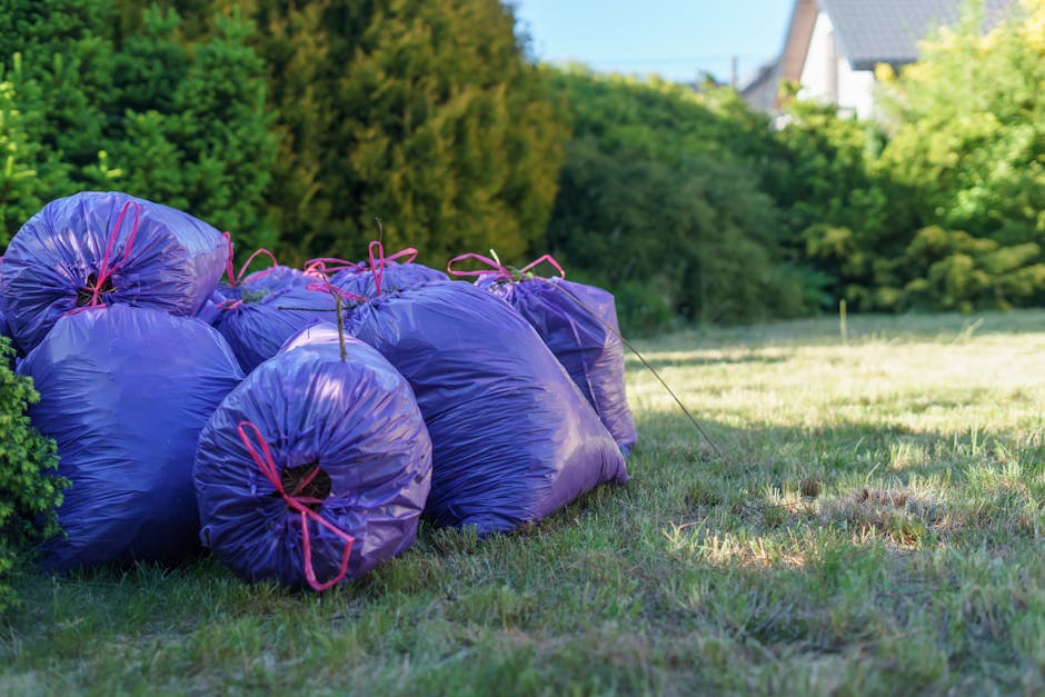 A group of several large, purple rubbish bags made of glossy, durable plastic with a slightly crinkled texture are tightly packed and tied with pink drawstrings, resting on a patch of well-maintained grass in an outdoor setting. The bags are piled together near a green shrubbery on the left side of the image, with their contents bulging and unevenly shaped, indicating they are filled with household waste or garden debris. In the background, there is a lush row of trees and bushes with dense foliage, displaying various shades of green, along with a glimpse of residential buildings with pitched roofs partially visible at the upper right corner of the image. The scene is illuminated by natural daylight, casting soft shadows on the ground and highlighting the shiny surface of the bags. This visual context aligns with private waste disposal services providing on-site clearance, illustrating a typical waste collection scenario for rubbish removal in a residential environment, as managed by Waste Removal Harringay.