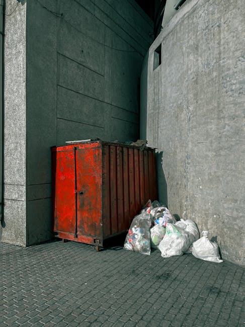A large, weathered red metal skip with visible rust and chipped paint is positioned against a building corner in an outdoor urban setting, with a small opening on one side. Surrounding the skip are several white plastic rubbish bags filled with waste, some of which are partially open, revealing mixed household rubbish inside. The skip and bags rest on a textured grey concrete pavement that extends into the background. The background features a dark green wall with a flat, smooth finish, and a light grey concrete wall with a slightly rough surface. The scene is illuminated by natural daylight, casting soft shadows, and highlights the contrast between the red, grey, and white tones, exemplifying typical private waste collection methods used for rubbish removal in tight street areas managed by independent services like Waste Removal Harringay.