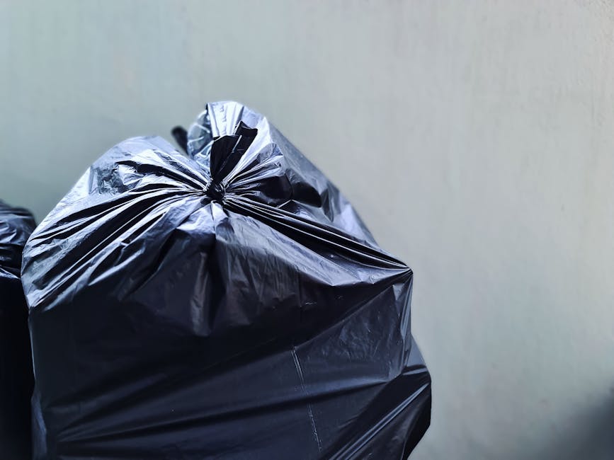 A group of several large, purple rubbish bags made of glossy, durable plastic with a slightly crinkled texture are tightly packed and tied with pink drawstrings, resting on a patch of well-maintained grass in an outdoor setting. The bags are piled together near a green shrubbery on the left side of the image, with their contents bulging and unevenly shaped, indicating they are filled with household waste or garden debris. In the background, there is a lush row of trees and bushes with dense foliage, displaying various shades of green, along with a glimpse of residential buildings with pitched roofs partially visible at the upper right corner of the image. The scene is illuminated by natural daylight, casting soft shadows on the ground and highlighting the shiny surface of the bags. This visual context aligns with private waste disposal services providing on-site clearance, illustrating a typical waste collection scenario for rubbish removal in a residential environment, as managed by Waste Removal Harringay.