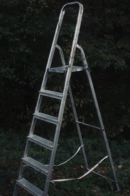 A silver, aluminum step ladder with four wide, textured steps, positioned outdoors on a grassy surface; the ladder's top platform is visible, and it leans slightly against a background of dense dark green foliage and shrubbery. The ladder has a safety strap attached to the lower rungs, indicating it may be used for safe access during on-site clearance or maintenance tasks. The environment appears natural, with diffuse lighting suggesting a cloudy day, and the scene is typical of a garden or backyard area where rubbish or waste materials might be temporarily placed before disposal. This scene highlights the type of equipment sometimes used alongside private rubbish removal services like those provided by Waste Removal Harringay for organizing or reaching higher areas during waste collection or refurbishment tasks, aligning with the theme of alternative waste handling methods outside standard council services.