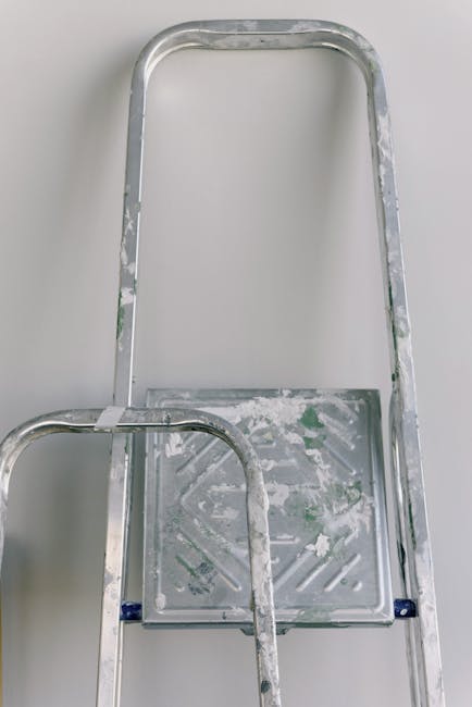 A close-up view of a silver, lightweight aluminium scaffold tower with a small platform, positioned on a plain white surface. The platform shows signs of wear with residual paint splatters and smudges in green and white. The structure's vertical and horizontal bars are visible, with some areas of minor scuffing and corrosion. The background is neutral and uncluttered, emphasizing the scaffold's utilitarian purpose. This equipment is often used for on-site clearance or access during rubbish removal projects, with Waste Removal Harringay offering alternative waste handling options outside of municipal collection services. The scene reflects a typical setup for private or professional waste clearance tasks, supporting the process of preparing debris or materials for disposal or recycling at a location such as a residential or commercial property in the Harringay area.