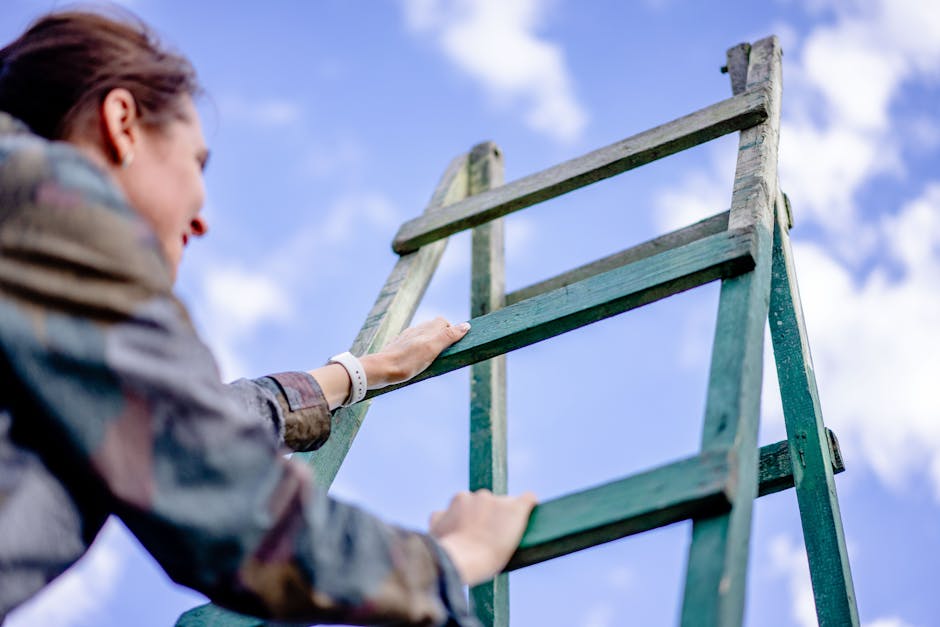 A silver, aluminum step ladder with four wide, textured steps, positioned outdoors on a grassy surface; the ladder's top platform is visible, and it leans slightly against a background of dense dark green foliage and shrubbery. The ladder has a safety strap attached to the lower rungs, indicating it may be used for safe access during on-site clearance or maintenance tasks. The environment appears natural, with diffuse lighting suggesting a cloudy day, and the scene is typical of a garden or backyard area where rubbish or waste materials might be temporarily placed before disposal. This scene highlights the type of equipment sometimes used alongside private rubbish removal services like those provided by Waste Removal Harringay for organizing or reaching higher areas during waste collection or refurbishment tasks, aligning with the theme of alternative waste handling methods outside standard council services.