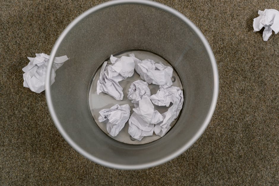 An overhead view of a cylindrical metal waste bin placed on a brown textured carpet. Inside the bin, there are several crumpled sheets of white paper, some submerged in a small amount of water at the bottom of the bin. Additional crumpled paper pieces are scattered on the floor around the bin, indicating a recent or ongoing rubbish collection process. The metallic surface of the bin is smooth and slightly reflective, contrasting with the rougher, fibrous texture of the paper and the coarse carpet. The environment appears to be an indoor setting, possibly an office space, relevant to areas where private rubbish removal or on-site waste clearance might be carried out by services such as Waste Removal Harringay, supporting the context of efficient rubbish disposal for local businesses.