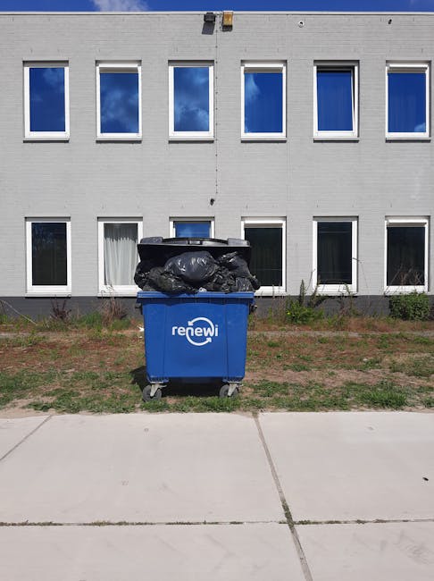 A person standing outdoors on a grassy area, wearing a plaid yellow and gray shirt over a green top, with gray trousers and a brown belt. They are wearing bright green gloves and holding open a large black rubbish bag made of thick, shiny plastic material, which is partially lined with a silver inner layer. The scene suggests the person is engaged in waste collection or rubbish removal, with the background showing a well-maintained lawn or garden. The natural lighting indicates daytime, and the environment appears clean and orderly, fitting within the context of independent rubbish disposal or on-site waste clearance facilitated by Waste Removal Harringay.