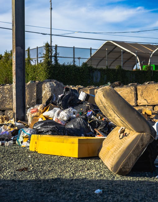 A close-up view of a pavement area with a pile of mixed waste, including black and clear plastic rubbish bags, discarded cardboard boxes, and various small debris, all casually dumped on the gritty, asphalt surface. An old, beige car tyre leans on the pile, partially covering some of the waste. Behind this clutter, there is a low stone wall made from roughly cut sandstone blocks, separating the waste from a background of green foliage and trees. To the left, a metal utility pole is visible, with electrical wires stretching across the scene under a partly cloudy blue sky, suggesting an outdoor, urban environment. The waste accumulation appears unmanaged, potentially indicative of an alternative private collection or on-site clearance scenario, serving as a practical example of rubbish that Waste Removal Harringay could assist with in compliance with local kerbside rules and regulations.