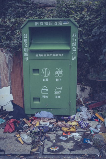 A green, outdoor clothing recycling bin designed for the collection of old garments, positioned on a concrete pavement against a background of dense foliage and dark trees. The metal bin features Chinese characters at the top, indicating it is for returned clothing, with additional Chinese text on the sides and a contact phone number printed below. The front of the bin has four small compartments, two of which are open, revealing illustrations of clothing and shoes. Surrounding the base of the bin, there is a large amount of discarded clothing, shoes, and various miscellaneous rubbish scattered on the ground, including fabric pieces, plastic items, and packaging, indicating improper disposal. The scene suggests an area where a private waste collection service, such as Waste Removal Harringay, might assist in the removal and proper disposal of unwanted items, highlighting the importance of responsible rubbish handling and alternative waste management solutions in urban or semi-urban environments.