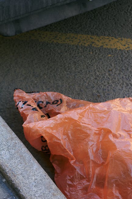 A close-up view of a pavement area with a pile of mixed waste, including black and clear plastic rubbish bags, discarded cardboard boxes, and various small debris, all casually dumped on the gritty, asphalt surface. An old, beige car tyre leans on the pile, partially covering some of the waste. Behind this clutter, there is a low stone wall made from roughly cut sandstone blocks, separating the waste from a background of green foliage and trees. To the left, a metal utility pole is visible, with electrical wires stretching across the scene under a partly cloudy blue sky, suggesting an outdoor, urban environment. The waste accumulation appears unmanaged, potentially indicative of an alternative private collection or on-site clearance scenario, serving as a practical example of rubbish that Waste Removal Harringay could assist with in compliance with local kerbside rules and regulations.
