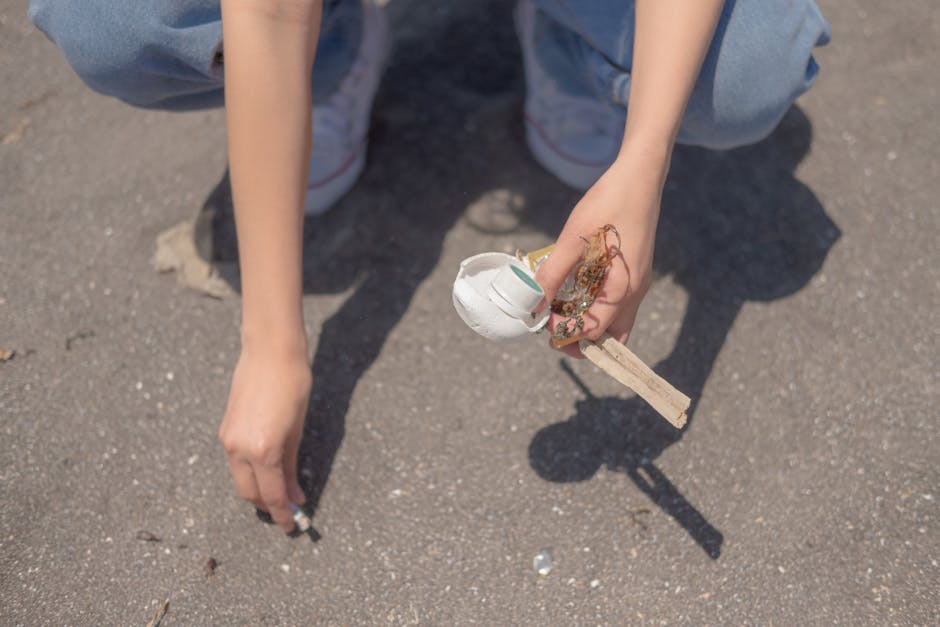 A person crouching on a paved surface, holding a small white plastic cup with a torn piece of wood and tangled string or twine in their right hand, while their left hand rests on the ground. The individual is wearing light blue jeans and white sneakers with a pinkish stripe, and the scene appears to be outdoors in natural daylight. The background reveals a rough, textured asphalt surface, with minor debris scattered around. This scene represents a typical instance of improper disposal or littering, which could be addressed through waste removal services such as those provided by Waste Removal Harringay, especially in contexts involving small plastics and mixed refuse often found in urban environments.