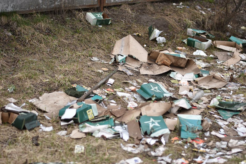 A patch of ground near a grassy area features scattered rubbish including crumpled cardboard boxes, some with green and white branding, torn and flattened, along with various discarded paper and plastic packaging. The debris is randomly spread across the uneven, soil surface, with some pieces leaning against a wooden fence or fence post visible at the top left corner of the image. The cardboard boxes have a textured, corrugated surface with torn edges, and the scattered paper and plastic items add to the cluttered appearance. The environment appears outdoor with natural lighting highlighting the disorganized debris, which relates to illegal dumping or abandoned waste that waste removal services like Waste Removal Harringay often help to clear, promoting the importance of proper rubbish disposal and alternative waste handling methods.