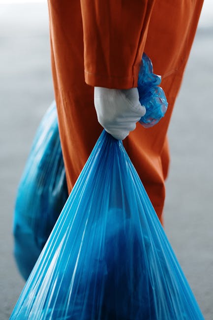 A person wearing an orange coverall and a white glove is holding the handles of a large blue plastic rubbish bag, which appears to be filled with waste material. The bag is conical and semi-transparent, showing the dark contents inside, and is gathered at the top, secured with a tied opening. The background is out of focus, suggesting an outdoor setting with neutral lighting. The individual is positioned with only their hand and part of their midsection visible, indicating they are engaged in rubbish collection or disposal activity. This scene exemplifies private waste handling and on-site rubbish removal efforts, similar to what services like Waste Removal Harringay offer by providing independent collection solutions outside of local authority systems. The surrounding environment, possibly a driveway or outdoor area, hints at a clean-up operation, supporting the context of professional rubbish removal or waste management services targeting domestic or commercial sites.