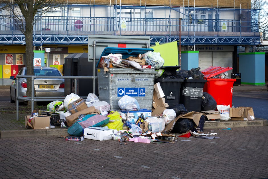 A collection of overflowing rubbish bins and scattered waste materials on a paved sidewalk in an urban area. The scene includes a large grey mixed paper and cardboard recycling bin that is overflowing with crumpled paper, cardboard boxes, and miscellaneous packaging, with some items spilling onto the ground. To the right, there are several black trash bags and a bright red waste bin containing additional rubbish. Surrounding the bins, various discarded items such as boxes, plastic containers, and paper debris are strewn across the pavement. In the background, there is a multi-storey building with scaffolding and construction barriers, indicating nearby ongoing construction work. A parked vehicle is visible behind a metal railing on the left side of the image, and a row of commercial storefronts with signage appears further back. The overall scene depicts a typical urban rubbish accumulation, highlighting the importance of waste management services like those provided by Waste Removal Harringay for alternative collection and on-site clearance options.